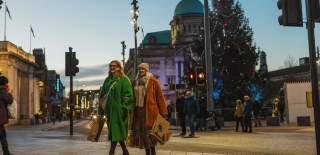 Two people wearing colourful winter coats walk arm-in-arm with shopping bags in Hull city street at dusk, passing a decorated Christmas tree and historic buildings in the background.