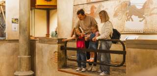 A family with two young children stands in the Hull & East Riding Museum, looking over a railing at an exhibit, with a large ancient mural depicting horses and chariots on the wall behind them.