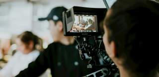A man looks through a filming camera in the foreground as the people being filmed are blurred in the background.