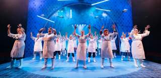 A stage scene bathed in blue light with women dressed in white aprons and wellington boots, their gloved hands raised up as they dance.