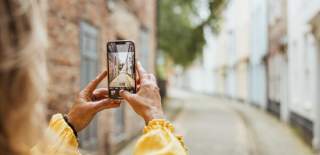 Prince Street in Hull's Old Town. The colourful, curving buildings are out of focus. A blonde woman stands with her back to the camera, holding her phone to take a photograph of the cobbled street. She is wearing an eye catching, flowy yellow top.