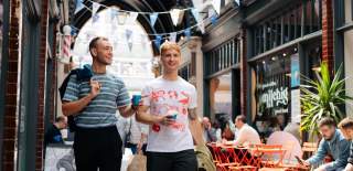 Two man strolling Paragon Arcade on a summer's day. The are holing takeaway coffees and smiling. The man on the left is wearing a blue striped t-shirt and black trousers with a dark jacket over his shoulder. The man on the right is wearing a white t-shirt with neon prints of animals on and dark trousers. Blue bunting is hung from the beamed ceilings. People are sat outside a café with orange seating and plants.