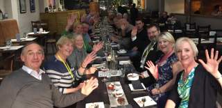 Large group seated together at Ambiente Hull,  raising hands in greeting, with plates, drinks and table settings laid out during a group dining event.