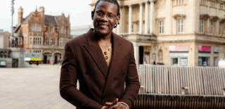 A man wearing glasses and a brown double-breasted coat smiles while standing in an open urban square with historic buildings in the background.