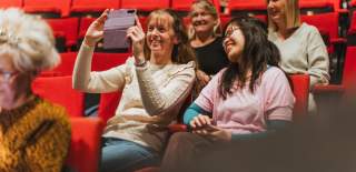 The audience at Hull Truck Theatre are seated on red chairs and the four ladies in the foreground are smiling and taking a selfie on a purple phone ready for the show to begin.