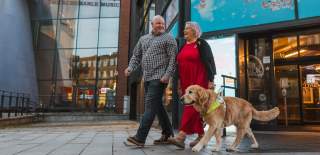 A man and woman walking with a guide dog along a city street outside Hull Truck Theatre.
