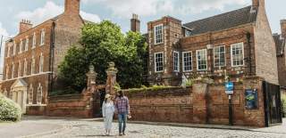 A cobbled street scene outside a historic red‑brick building in Hull’s Museums Quarter, with two visitors walking past the Wilberfoce House under a bright, clear sky.