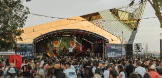 The crowd at Humber Street Sesh at sunset. The crowd are wearing summer clothes and are watching a band at the Hull Trains stage which is branded in the blue Hull Trains logo with hearts. Behind the stage, The Deep's iconic sloping arcitecture shines in the sunset.