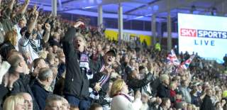 A packed football stadium with fans standing shoulder to shoulder, cheering and clapping with raised arms. Many wear scarves and team colours, some wave Union Jack flags, and a large screen on the right displays “Sky Sports Live” as the crowd celebrates mid-match.