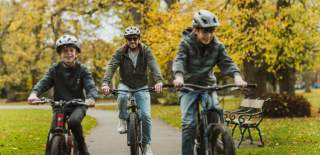 Three cyclists riding along a tree‑lined path in Hull’s Pearson Park during autumn, with fallen leaves and park benches visible