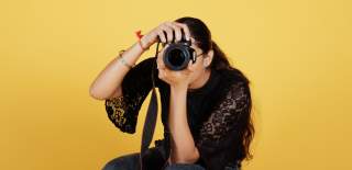 A woman holds a camera up covering her face in front of a yellow background