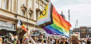 Pride In Hull's parade walking down Carr Lane. The sun is shining, the crowd is smiling and dressed in shorts and t-shirts. A group of people holding a rainbow Smith+Nephew banner are in the foreground with a brightly dressed woman waving an Intersex Inclusive pride flag. Many other pride flags can be seen in the background.