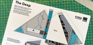 A printed cut‑out model template of The Deep aquarium in Hull laid on a green cutting mat, showing labelled sections and assembly instructions alongside a metal ruler.