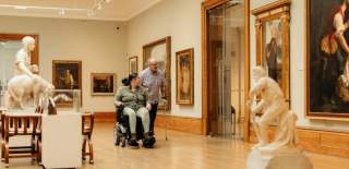 A wheelchair user explores Ferens art gallery Hull with another visitor, surrounded by sculptures and framed artwork. The scene highlights inclusive museum accessibility