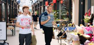 Two people walking through a bright indoor arcade lined with independent shops and colourful flower displays, browsing retail stalls in Hull’s Paragon Arcade.