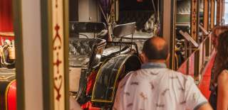Visitors viewing vintage vehicles and historic carriages inside a museum gallery, featuring ornate display panels and classic leather seating on the exhibits