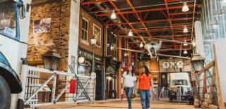 The main concourse of Streetlife Museum. The beamed ceilings are lied with bright lights and a propellor plane hangs from the rafter. Below is a replica 1950s high street. A vintage OXOsign is on the back wall with an retro van parked in front. Two women are walking underneath the plan across an old train cross towards the camera. They are looking up at the exhibits and are wearing t-shirts and jeans.