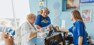 A staff member at a visitor attraction reception desk assisting two customers with tickets, highlighting accessible customer service and tourism support in a bright, welcoming environment.