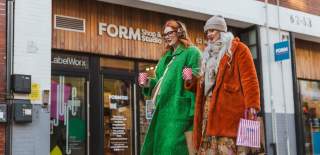Two women walk down Humber Street's cobbles. They stroll past FORM Shop's wooden exterior with a colourful display in the window. They are bundled up in hats and scarves and are carrying coffees and shopping bags.