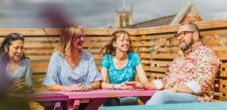 A man and woman  wearing a hearing aid relaxes group of friends at a colourful outdoor seating area on Humber Street in Hull. The scene showcases the vibrant, accessible social spaces of the Humber Street area