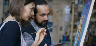 Two artists stand in front of an easel gazing at the painting, mid discussion.