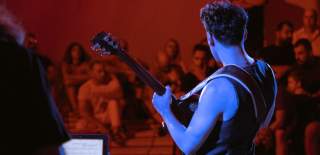 A man plays an electric guitar to an audience. He faces away from the camera and is illuminated by blue stagelights.