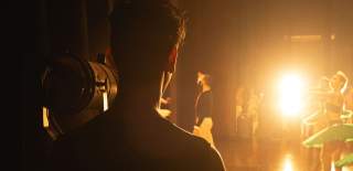 A man stands behind the curtains of a stage waiting to go on whilst ballet dancers are on stage. The spotlight shines towards the man as he faces with his back to the viewer.
