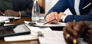 A close up of hands on a table during a meeting. There's paper, a water bottle, calculator and pens on the table.