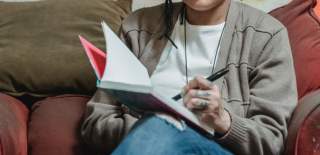 A woman sits on a sofa and writes in a notebook.
