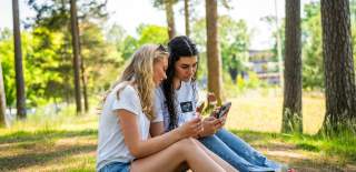 To jenter som sitter i parken og ser på telefonen til hverandre