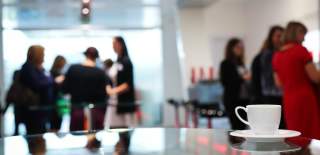 A photograph showing a cup and saucer in the foreground, with people in smart clothes in the background at a conference event