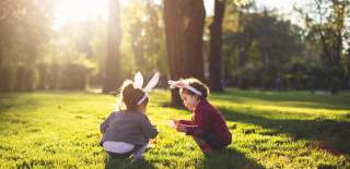 Two young children wearing bunny ears in a grassy field with the sun shining through the trees behind them