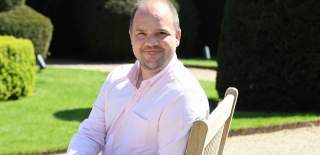 An image of Liam Bartlett, Chair of Shakespeare's England sitting in a chair outdoors in manicured gardens and smiling at the camera.