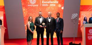 Representatives from Warwick Conferences pose on stage at the VisitEngland Awards for Excellence 2025–2026 ceremony, holding a tourism award in front of a brightly lit orange backdrop displaying the event branding. Four formally dressed attendees stand on a red carpet during the awards presentation.