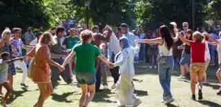 Group of people holding hands and dancing in a circle in Palmerston Park for Refugee Week