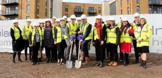 A group of people wearing hig vis and hard hats on a new development site. The two people in the middle have spades they are posing with for the camera