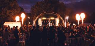 Nighttime outdoor concert at Mela Festival with a crowd watching performers on a lit stage, surrounded by fireworks and flames shooting into the sky.