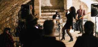 Band performing as per of music in the city inside an historic vault. There are two guitarists a drummer and a lead singing in a black dress, long curly hair and boots.