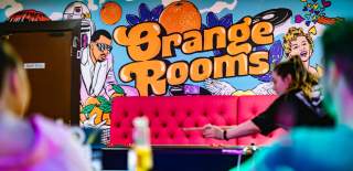Someone playing pool inside Orange Rooms with a bright pink sofa in the background and an Orange Rooms sign on the wall