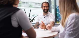 3 people sat round an office table talking and smiling. One person is facing the camera and has a white polo top on with their hands on the table.