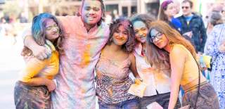 5 people with their arms around each other covered in colourful powder smiling at the Holi event in Guildhall Square
