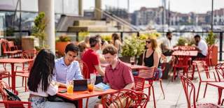 Red tables and chairs outside in Southampton with people sat round them. Three people in the foreground sat at a table looking at a laptop and smiling