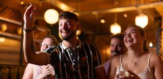 Group of friends smiling and laughing in a warmly lit bar while a man prepares to throw a dart.