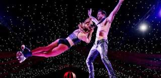 Two people performing an acrobatic stunt. One person is wearing just trousers and no top whilst someone else is hanging around their neck as they spin around.