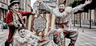 Three people dressed as clowns in grey and red clothing in front of a yellow and red sign that says Cirque du Kaka.