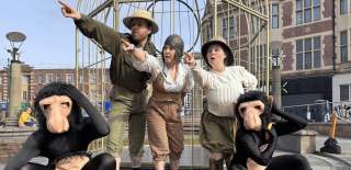 Five performers in a public square act out a scene in front of a large metal cage structure. Three in the centre wear vintage explorer-style clothing and point urgently into the distance with surprised expressions, while two others dressed in black bodysuits with ape masks crouch in the foreground, mimicking confused gestures.