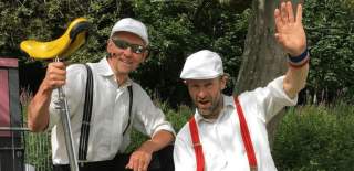 Two people wearing white shirts and braces. One is holding a unicycle and the other is waving at the camera.