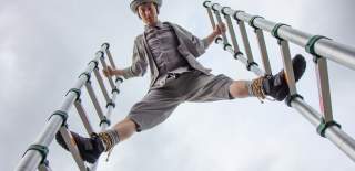 A performer dressed in grey with a grey hat on. They are standing between to ladders looking down at the camera.