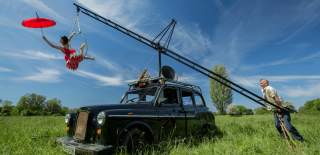 An old London taxi with a ladder on the top of it with two people either end. There is one person doing acrobatics off the higher end holding a red umbrella whilst a second person holds the other end of the ladder down to the floor.