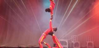 Three acrobats wearing red body suits. One is lying on the floor holding up another person with their legs. A third person is doing a hand stand on the second person's hands in the air.